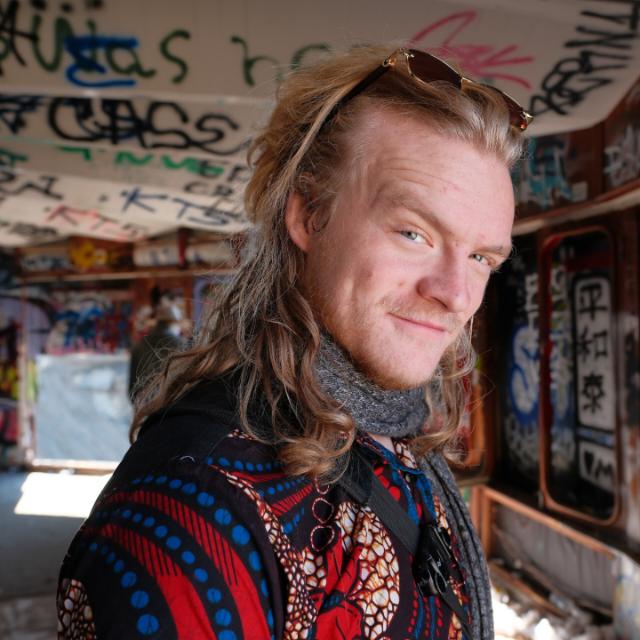 Bentley Moon: shoulder-length fair hair with sunglasses pushed up into it, a grey knitted scarf, and a red and blue patterned shirt, standing in a room with graffiti-covered walls and ceiling.